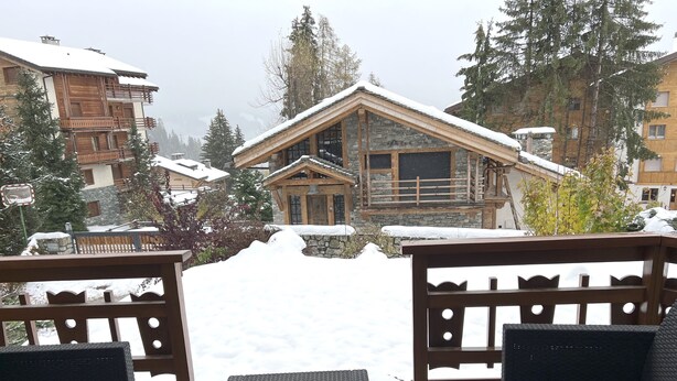 A wooden deck with wicker furniture overlooking a snowy landscape and a stone building with a sloped roof. - "Petit flocon" Apartment in the center of Verbier (Val de Bagnes)