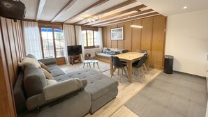 A modern living room with a grey sectional sofa, a wooden dining table with chairs, a TV on a stand, and large windows with curtains. - "Petit flocon" Apartment in the center of Verbier (Val de Bagnes)