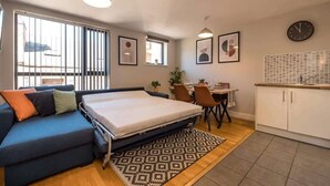 Une pièce de taille modeste comprenant un lit, un canapé, une table à manger avec des chaises, un coin cuisine et une fenêtre avec des stores.