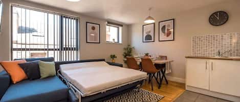 A compact room with a bed, a sofa, a dining table with chairs, a kitchen area, and a window with blinds.
