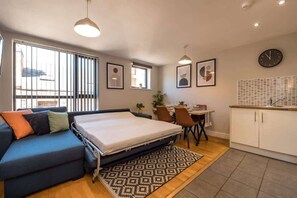 A compact room with a bed, a sofa, a dining table with chairs, a kitchen area, and a window with blinds.