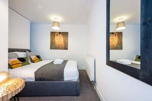 A hotel room with a bed, a bedside table, a mirror, and a painting on the wall. - Host & Stay | Preston's Rooftop Apt (Liverpool)