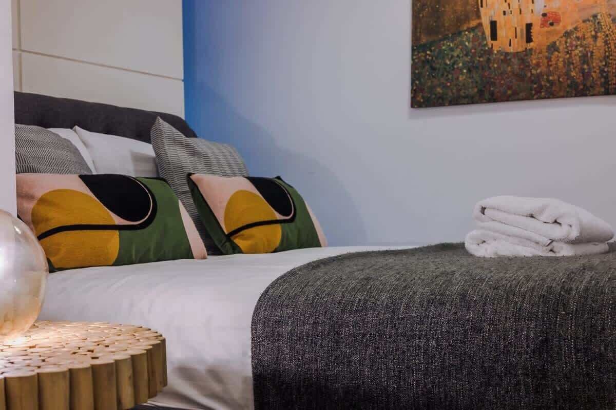 A neatly made bed with a grey blanket, a pillow with a yellow and green design, and a rolled white towel on the side.