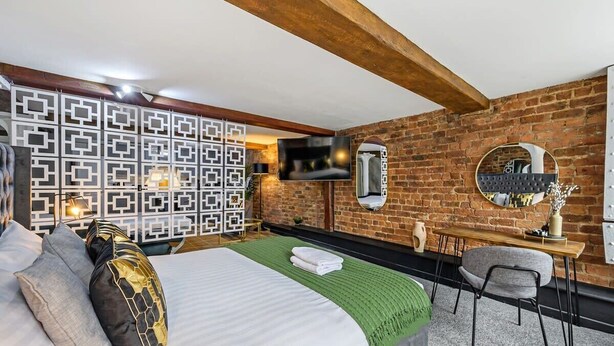 Habitación de hotel con pared de ladrillo, una cama con almohadas, un televisor, un espejo y un escritorio con una silla.