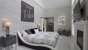 A modern bedroom with a curved bed, a dresser, a nightstand, and a fireplace.