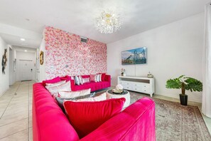 A living room with a vibrant pink sofa, a floral wallpaper, a white console table, and a painting on the wall. - Beautiful 2BR Condo w/ Private Balcony near Disney (Kissimmee)