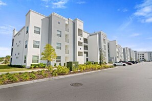 A modern multi-story residential building with a clean, white facade, a parking lot with several cars, and a clear blue sky. - Beautiful 2BR Condo w/ Private Balcony near Disney (Kissimmee)