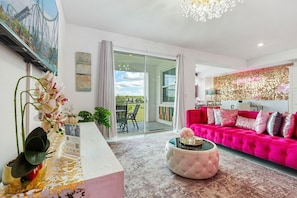 A modern living room with a pink sofa, a round coffee table, and a large window with a view of greenery. - Beautiful 2BR Condo w/ Private Balcony near Disney (Kissimmee)