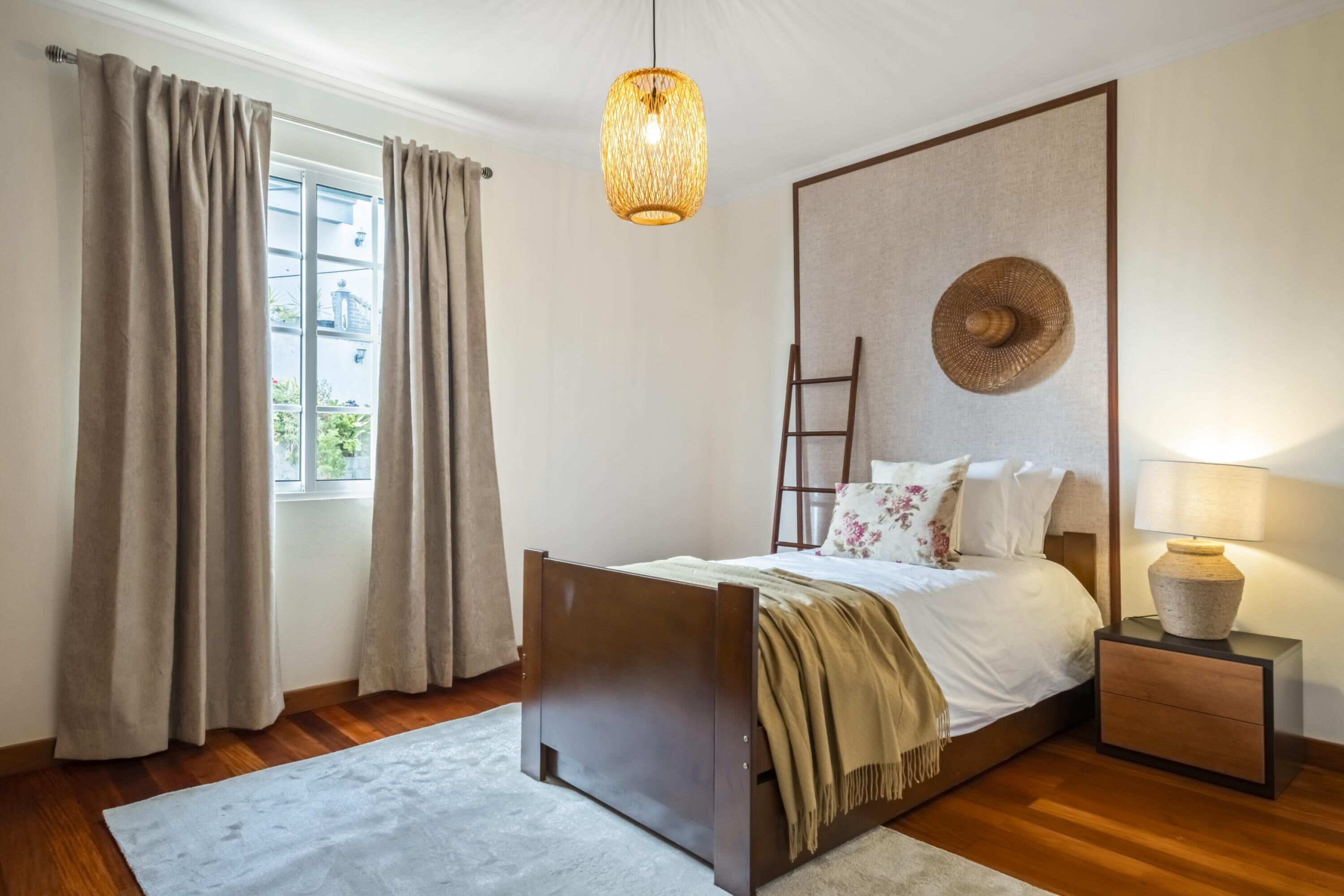 A bedroom with a bed, a nightstand, a lamp, a window with curtains, and a hanging light fixture.
