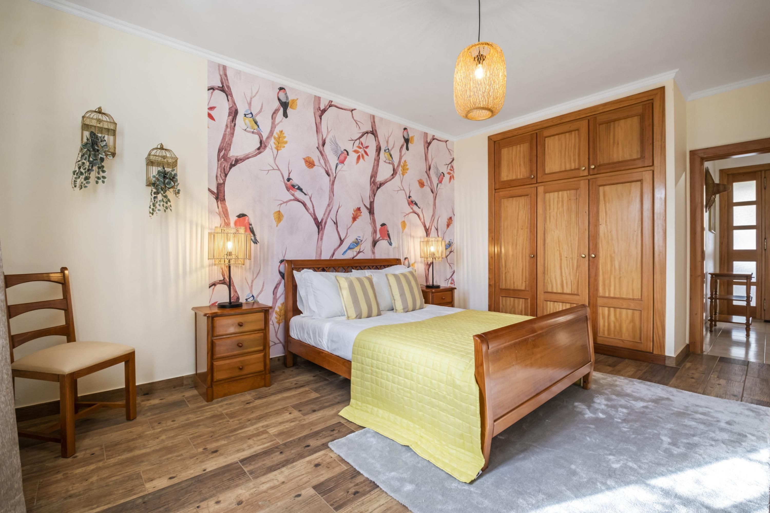 A bedroom with a wooden bed, a nightstand, a chair, a wardrobe, and a wall mural.