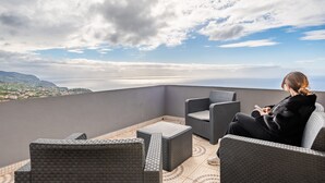 A balcony with wicker furniture, a view of the sea, and a person using a mobile device. - Sunny House by Homie (Ponta do Sol)