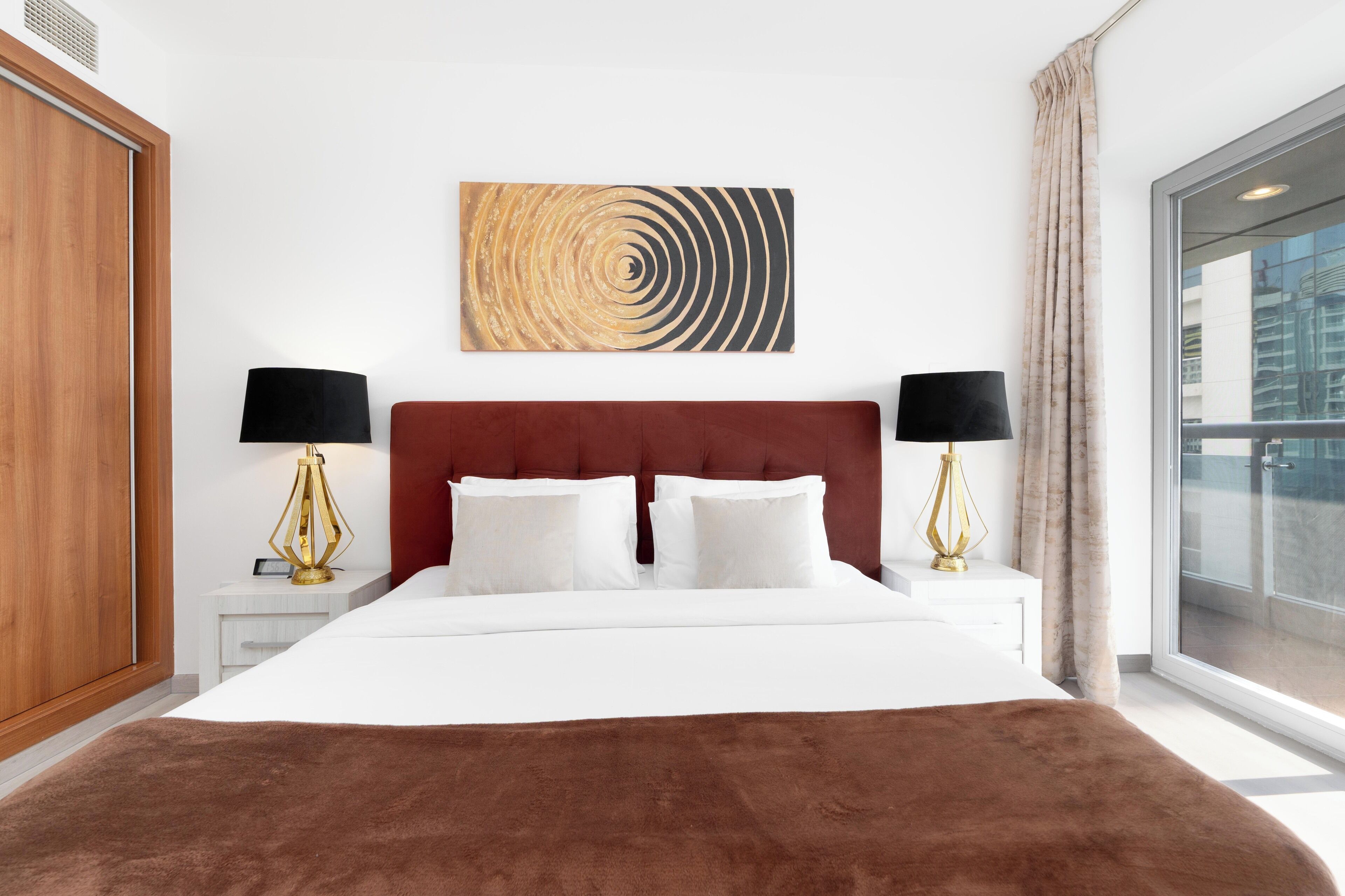 A hotel room with a large bed, two bedside tables with lamps, a wall art piece, and a large window with a view of buildings.