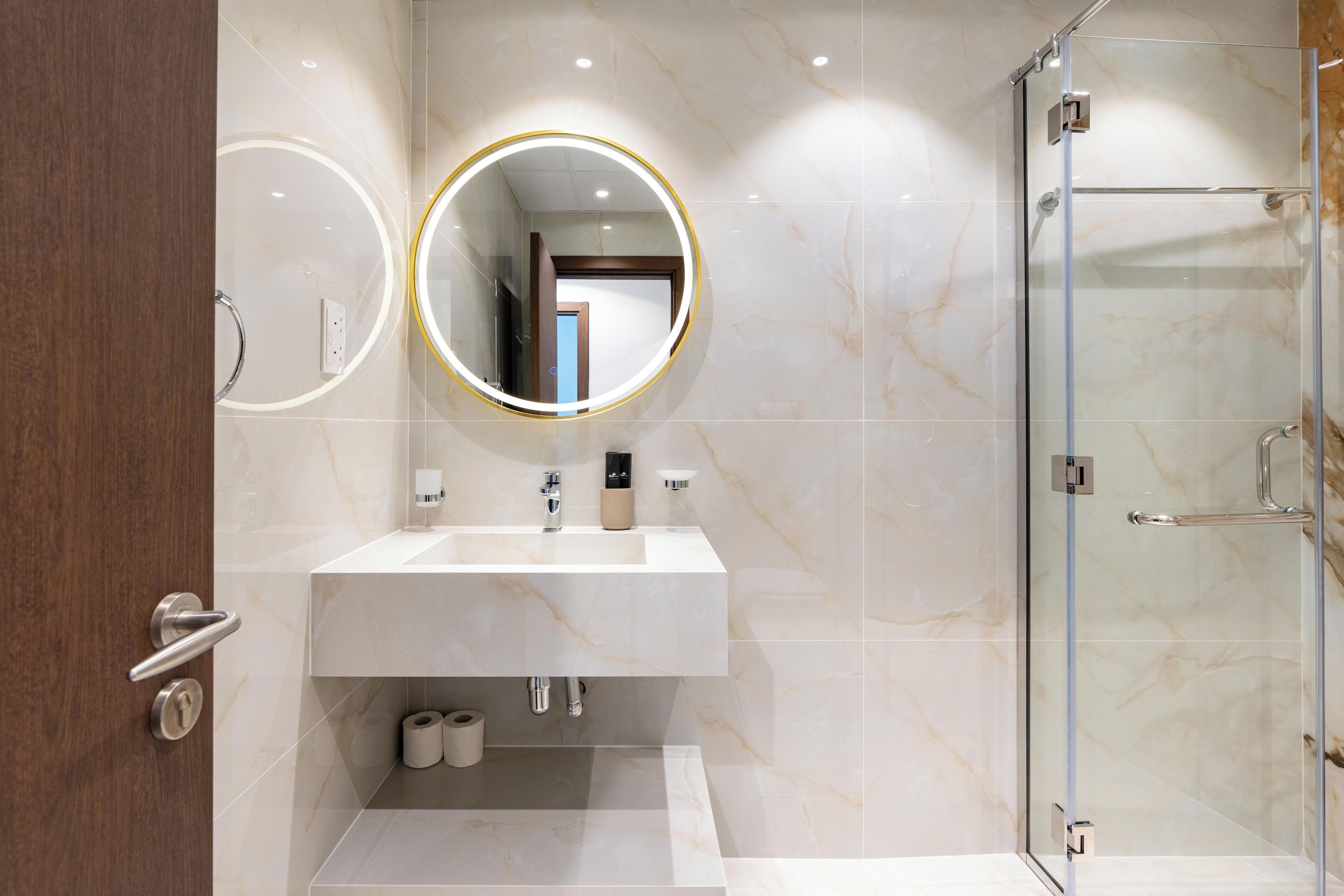 A modern bathroom with a marble sink, a round mirror with a gold frame, and a glass shower enclosure.
