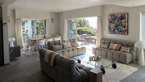 A spacious living room with a glass coffee table, grey sofas, and a dining area with a table and chairs.