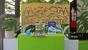 Un mostrador de recepción con una decoración verde y blanca, un letrero que dice 'J&L Homestay', un folleto de 'maya' y un letrero que indica 'CAJA DE PROPINA'.