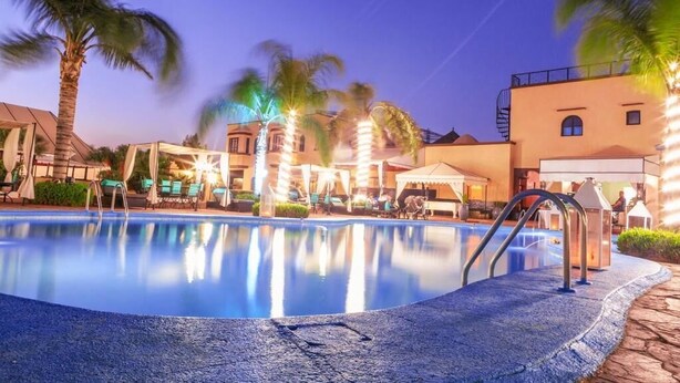 Ein Swimmingpool mit Liegestühlen und Sonnenschirmen, umgeben von Palmen und Gebäuden mit warmer Beleuchtung.