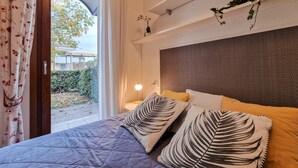 A bedroom with a bed, pillows, a nightstand, a lamp, and a window with curtains. - Comfortable Garden Apartment for Four Guests (San Michele al Tagliamento)