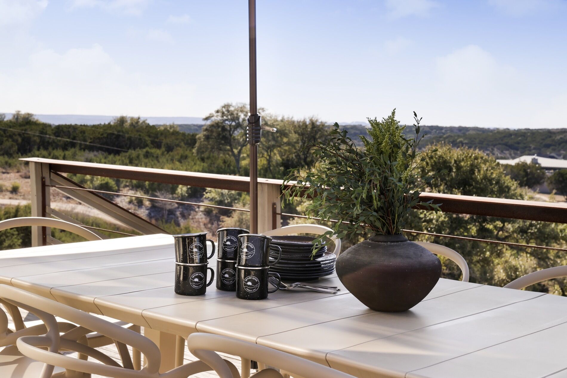 A table with a vase and black mugs on a balcony overlooking a scenic landscape.