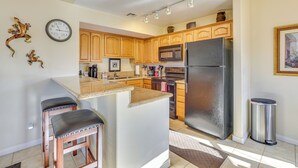 A kitchen with wooden cabinets, a stainless steel refrigerator, a microwave, and a sink. There are two bar stools and a clock on the wall. - Resort Pool & Spa Access: Mesquite Condo! (Mesquite)