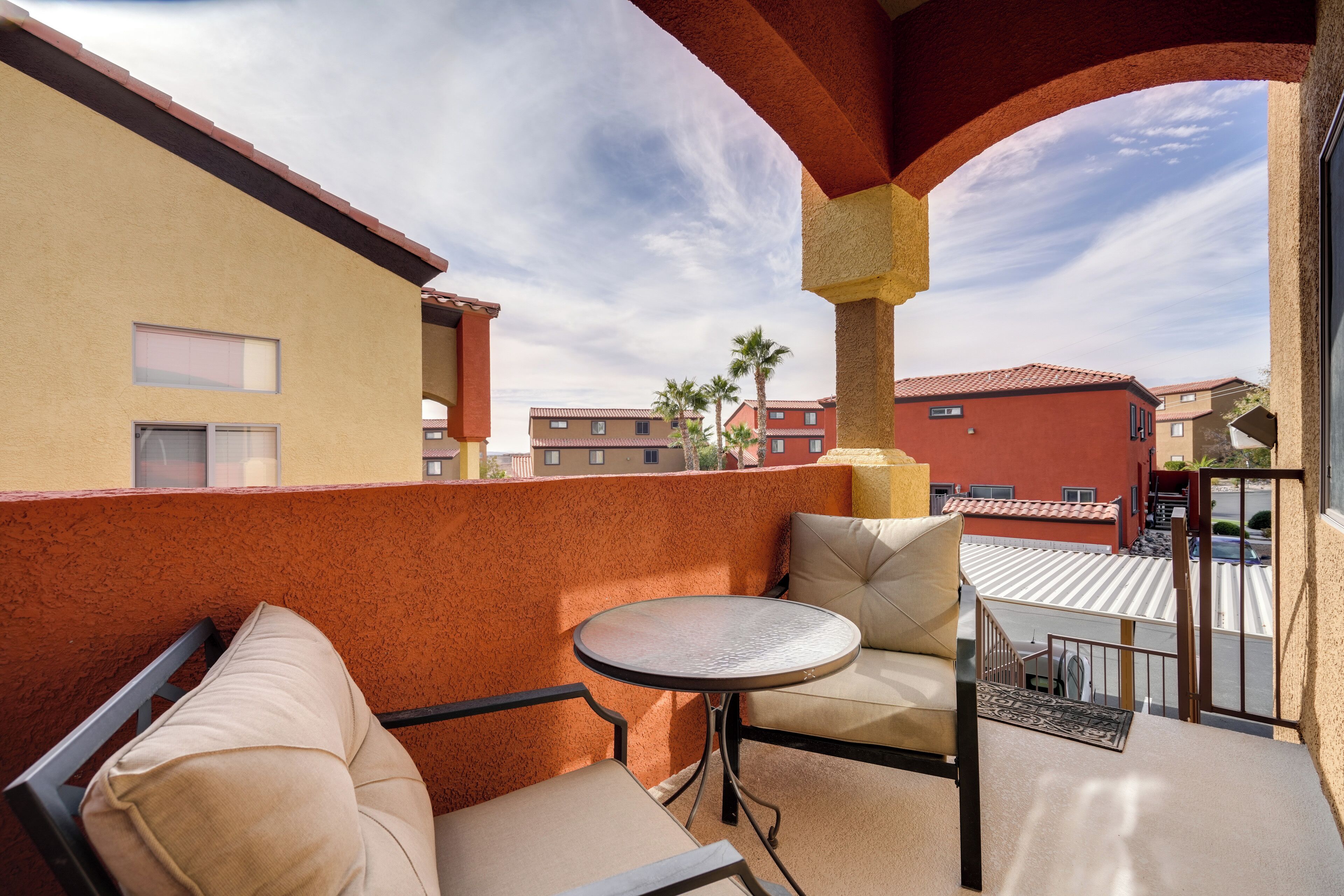 Een balkon met een ronde tafel en twee stoelen, met uitzicht op omliggende gebouwen en een heldere lucht.