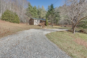 A gravel driveway leading to a single-story house with a porch, surrounded by trees and a grassy area.