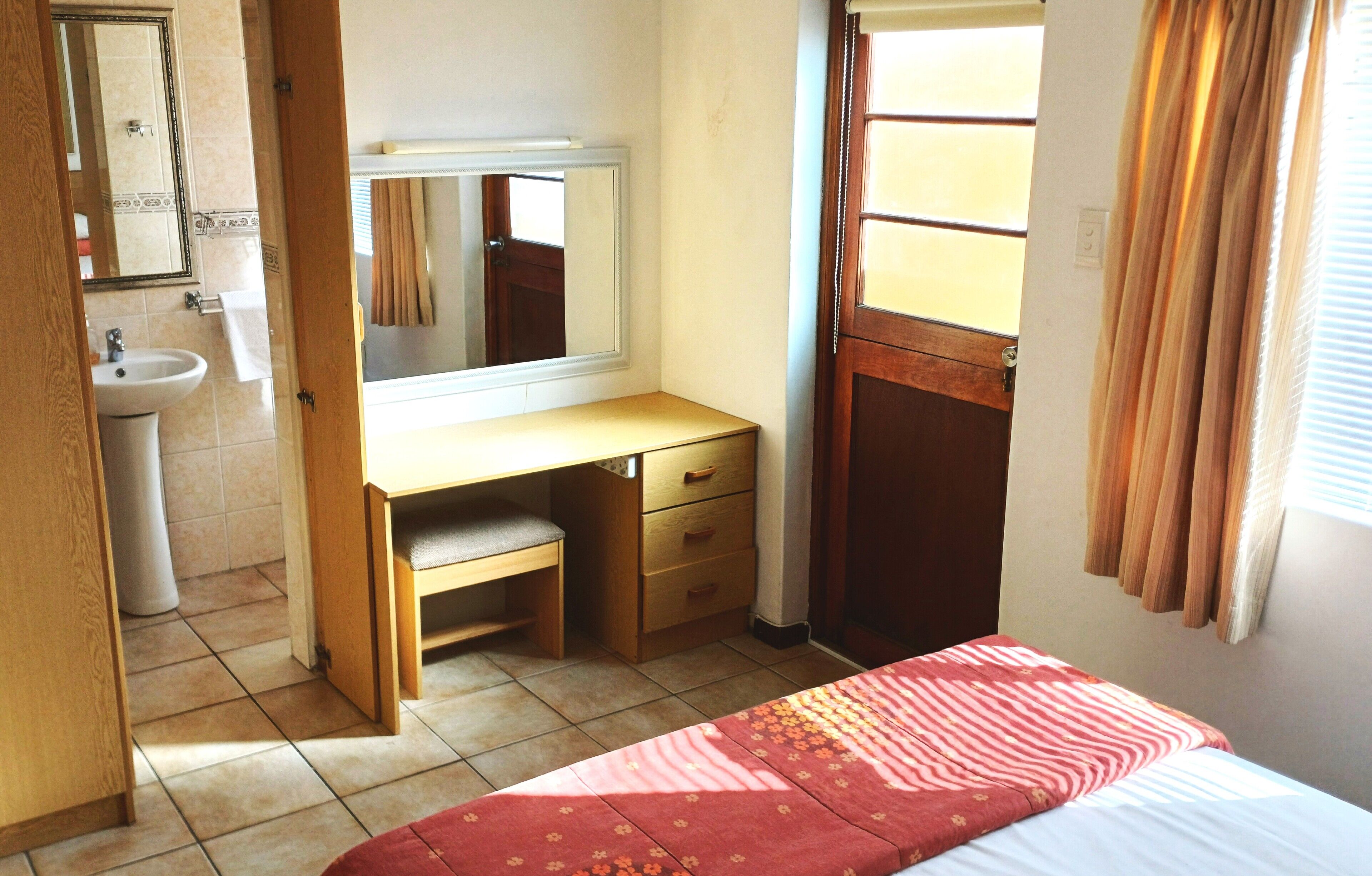 A bedroom with a bed, a wooden desk with a mirror, a sink, and a window with curtains.