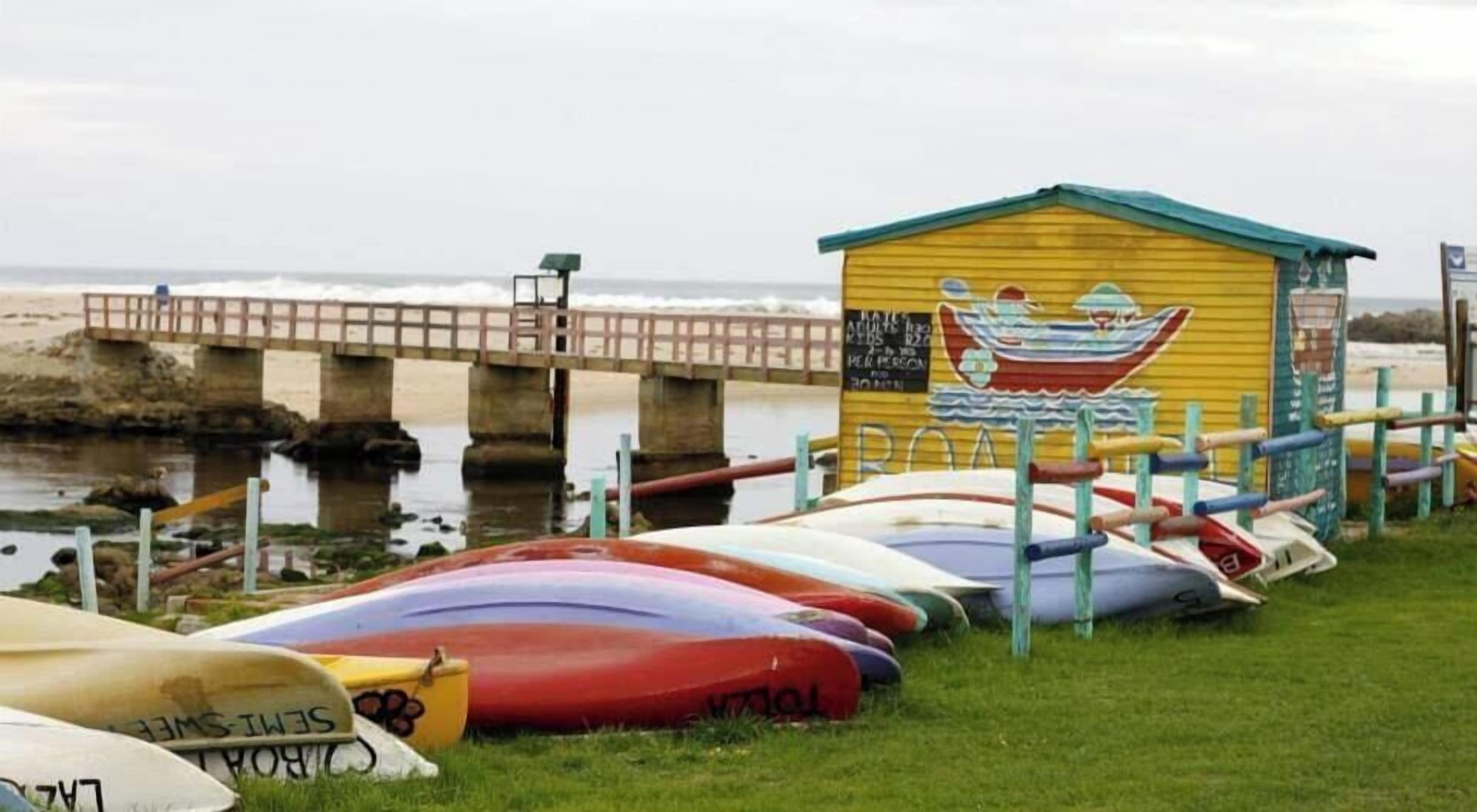 Una fila di kayak disposti su un'area verde con un piccolo edificio decorato da un murale a tema nautico.