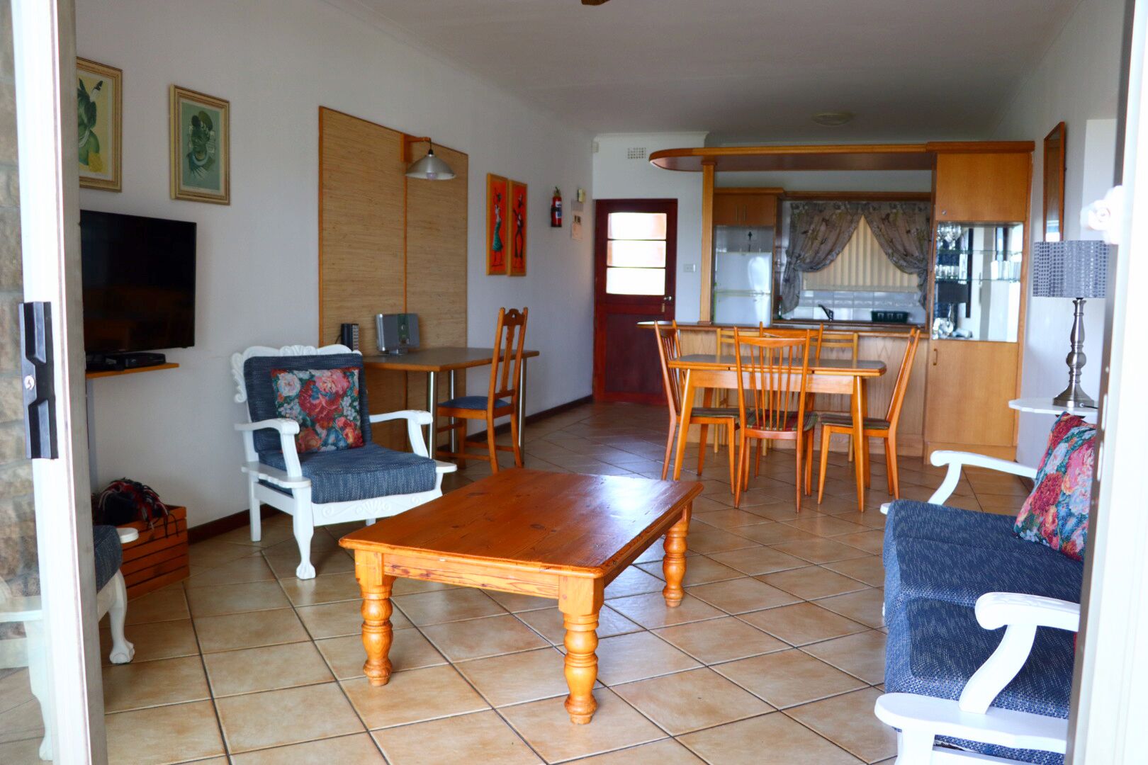 A living room with a wooden coffee table, two white armchairs with floral cushions, a TV, and a dining area with wooden chairs and a table.