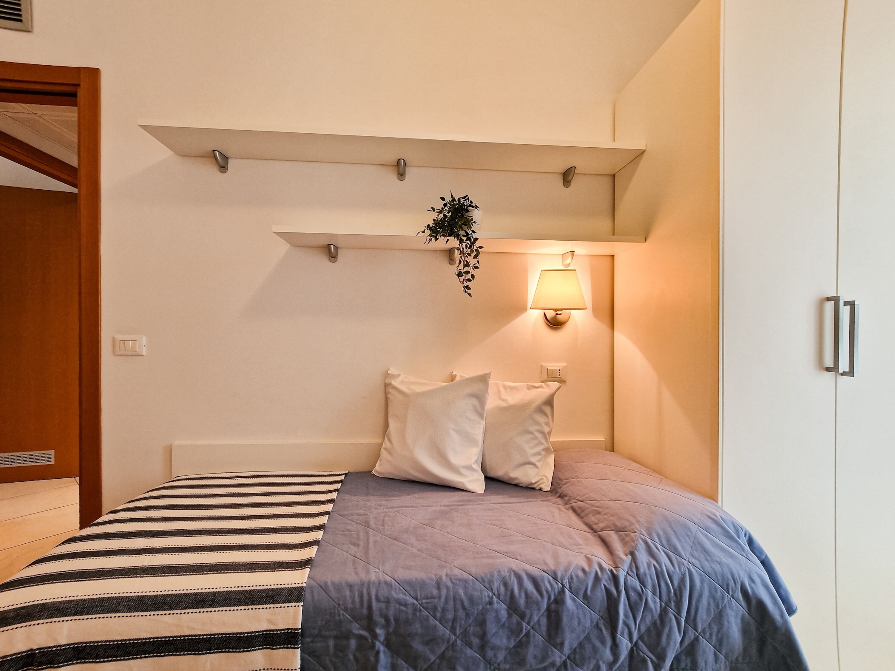 A bedroom with a bed, pillows, a bedside lamp, a shelf with a plant, and a wardrobe.