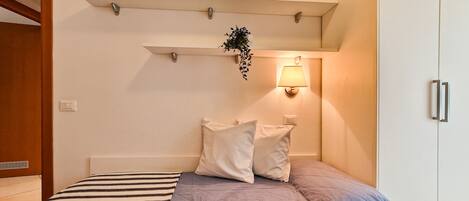 A bedroom with a bed, pillows, a bedside lamp, a shelf with a plant, and a wardrobe.