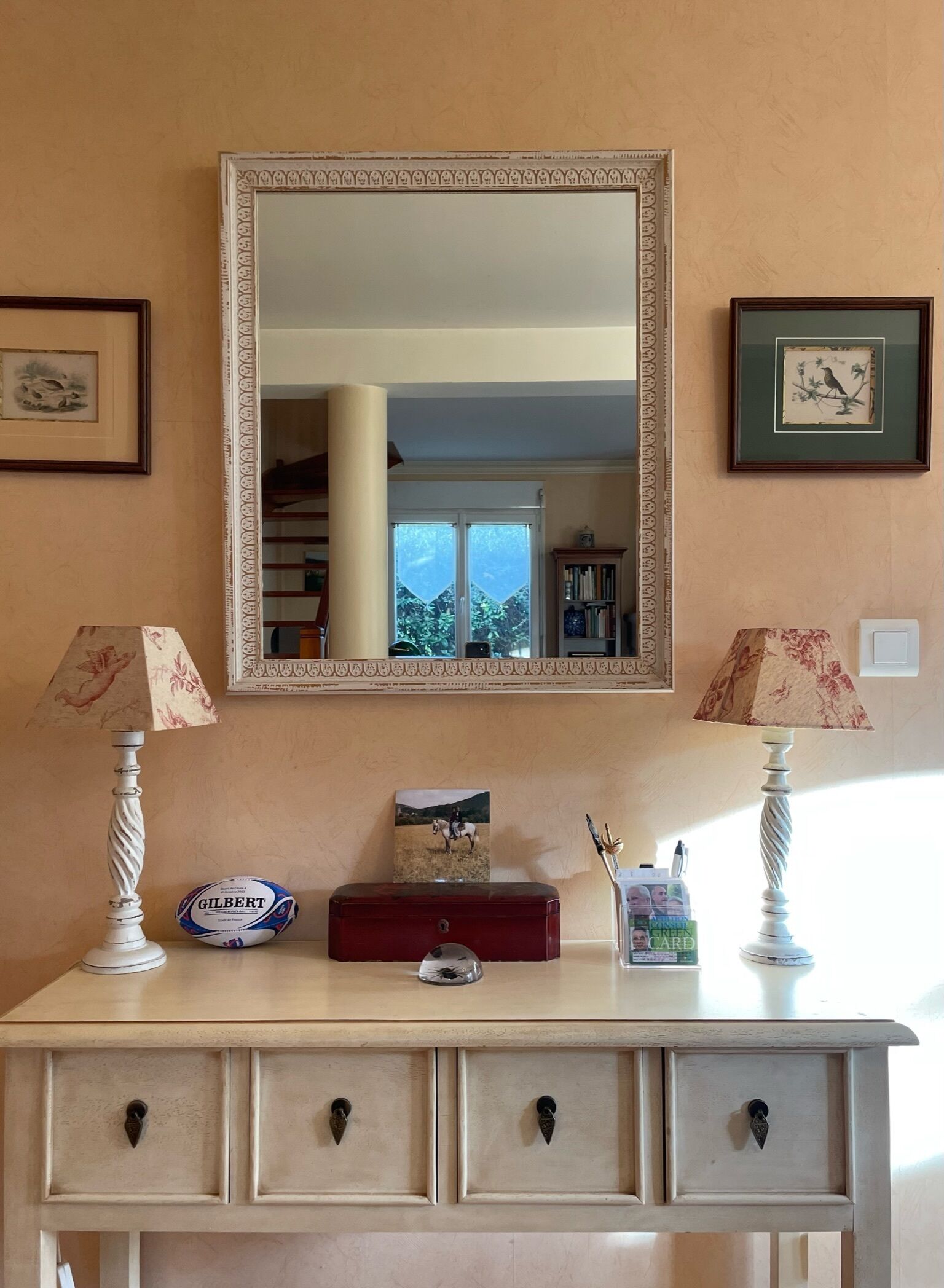 Habitació amb un mirall gran, dues làmpades, una taula petita amb una pilota de rugbi Gilbert, una targeta verda i una fotografia emmarcada d'un cavall.