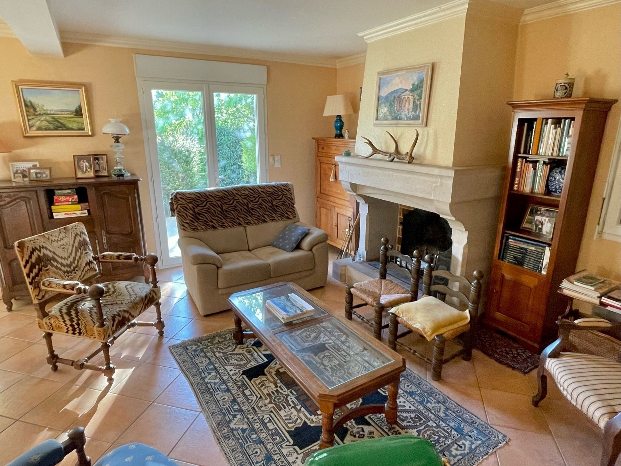 A living room with a sofa, armchairs, a fireplace, a bookshelf, and a large window.