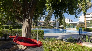 A pool area with a red swing chair, a palm tree, and a building in the background. - Palm Beach Sea View Apt by TrulyCyprus (Limassol)