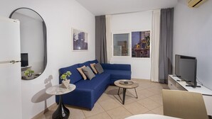 A modern living room with a blue sofa, a small round table, a flat-screen TV, and a large window with curtains. - Palm Beach Sea View Apt by TrulyCyprus (Limassol)