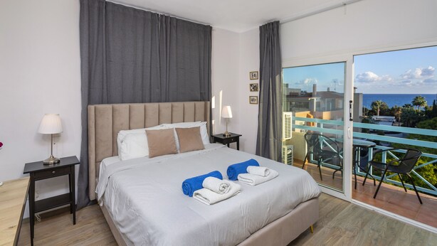 A hotel room with a large bed, two bedside tables, a nightstand, a balcony with a view of the sea, and a chair. - Palm Beach Sea View Apt by TrulyCyprus (Limassol)