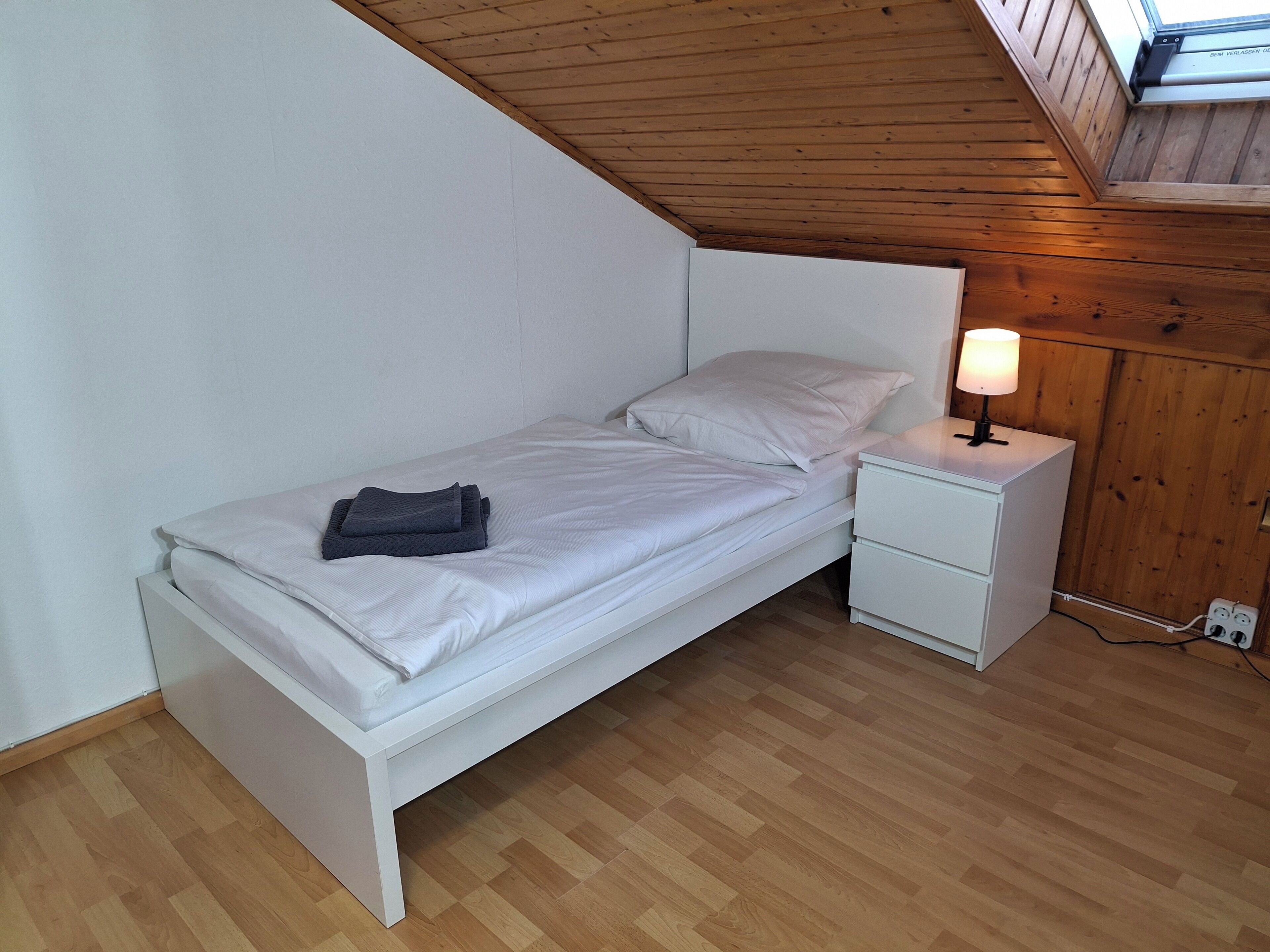 A single bed with white bedding, a bedside table with a lamp, and a wooden ceiling.