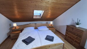 A bedroom with a wooden ceiling, a skylight, a bed with white linens, two bedside tables with lamps, and a wooden dresser.