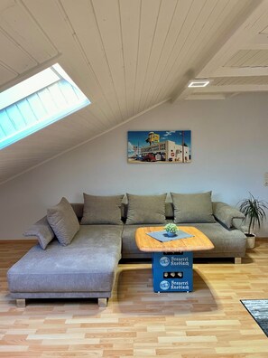 Uma sala de estar moderna com um sofá seccional, uma pequena mesa de centro de madeira, um vaso com planta e um quadro retratando uma cena de mercado.