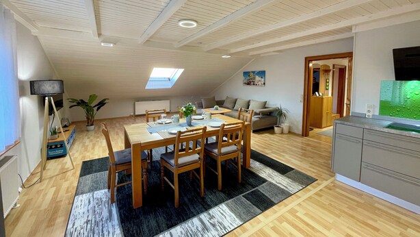 A spacious living area with a dining table, chairs, a sofa, a kitchen area, and a skylight.