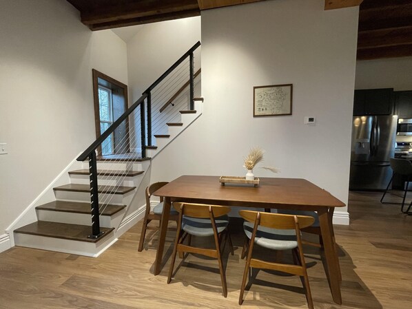 Uma área de refeições com uma mesa e cadeiras de madeira, uma escada com corrimão preto e uma cozinha ao fundo.