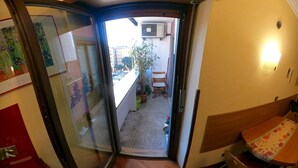 Um corredor estreito com porta que dá para uma varanda com vista para a rua. Há uma pequena mesa e cadeiras lá fora, e um vaso de planta na varanda.