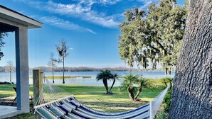 Eine Hängematze, die zwischen zwei Holzpfosten gespannt ist, eine Holzterrasse und der Blick auf einen See mit Bäumen und blauem Himmel.