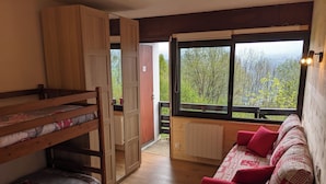 A cozy room with a bunk bed, a sofa with red cushions, and a view of greenery through large windows. - Duplex 4/6 Pers Dans une Ambiance Chaleureuse Avec vue Imprenable sur le Sancy (Murat-le-Quaire)