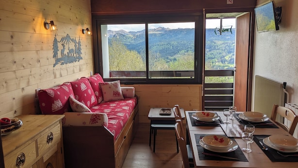 A cozy room with a wooden wall, a sofa with red cushions, a dining table set for two, and a large window offering a mountain view. - Duplex 4/6 Pers Dans une Ambiance Chaleureuse Avec vue Imprenable sur le Sancy (Murat-le-Quaire)