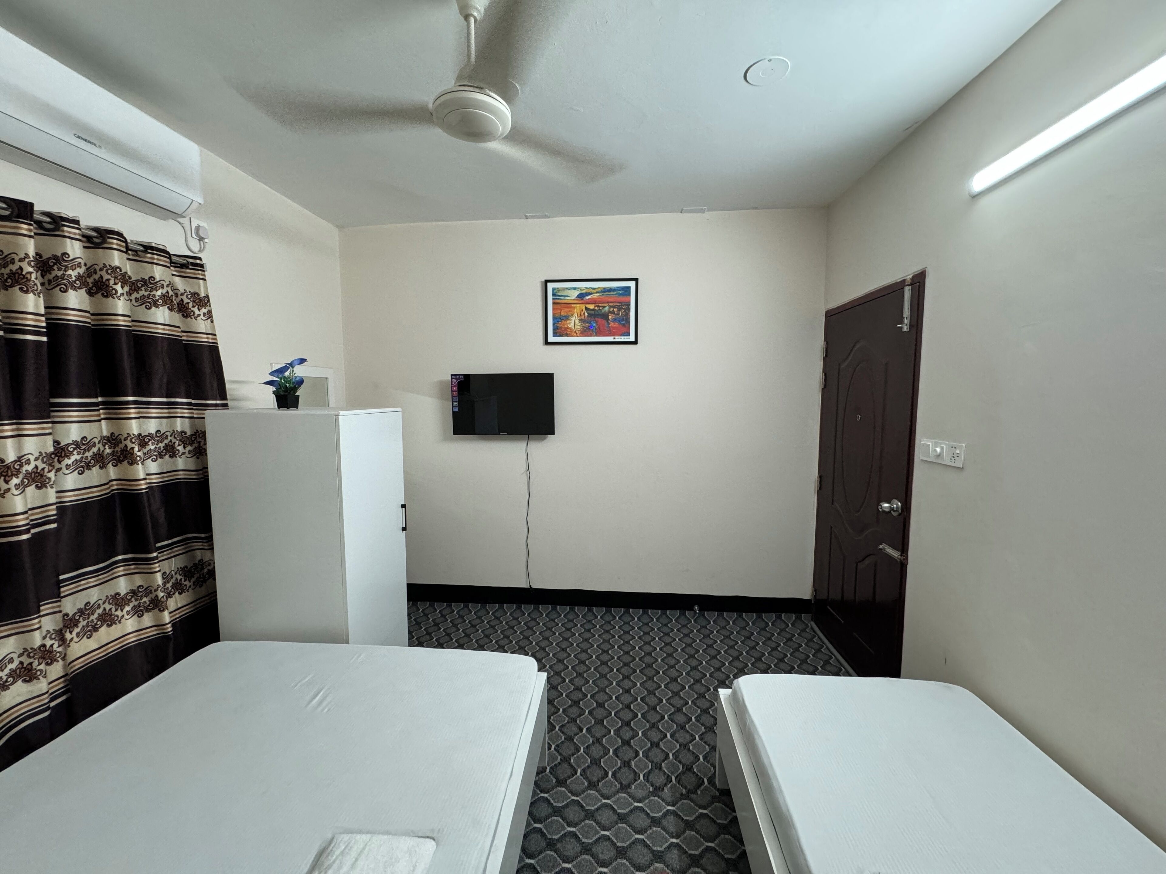 Une chambre d’hôtel avec deux lits, un ventilateur de plafond, un téléviseur à écran plat fixé au mur et une porte.