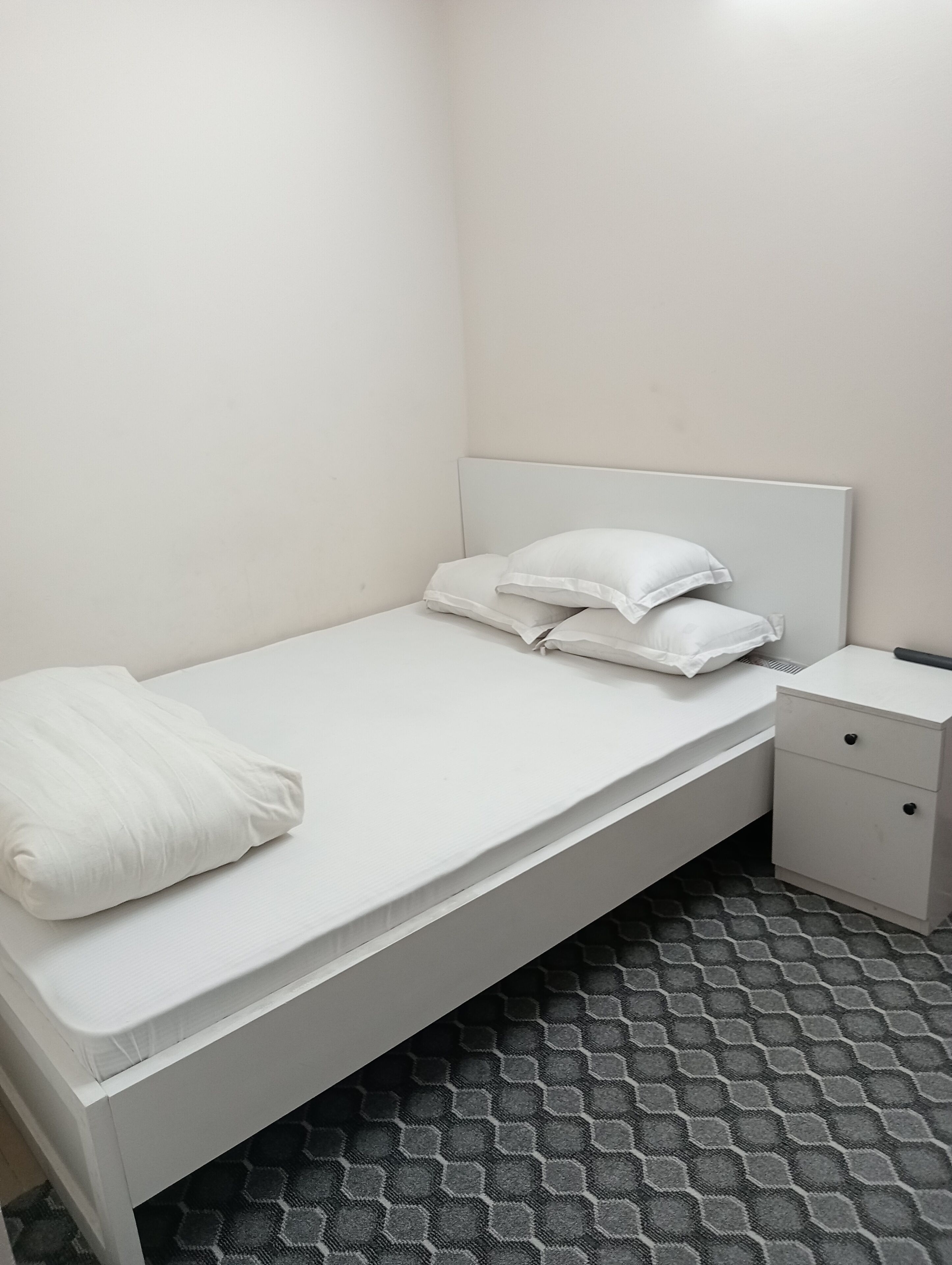 A small, single-bed room with a white bed, white nightstand, and a patterned floor.