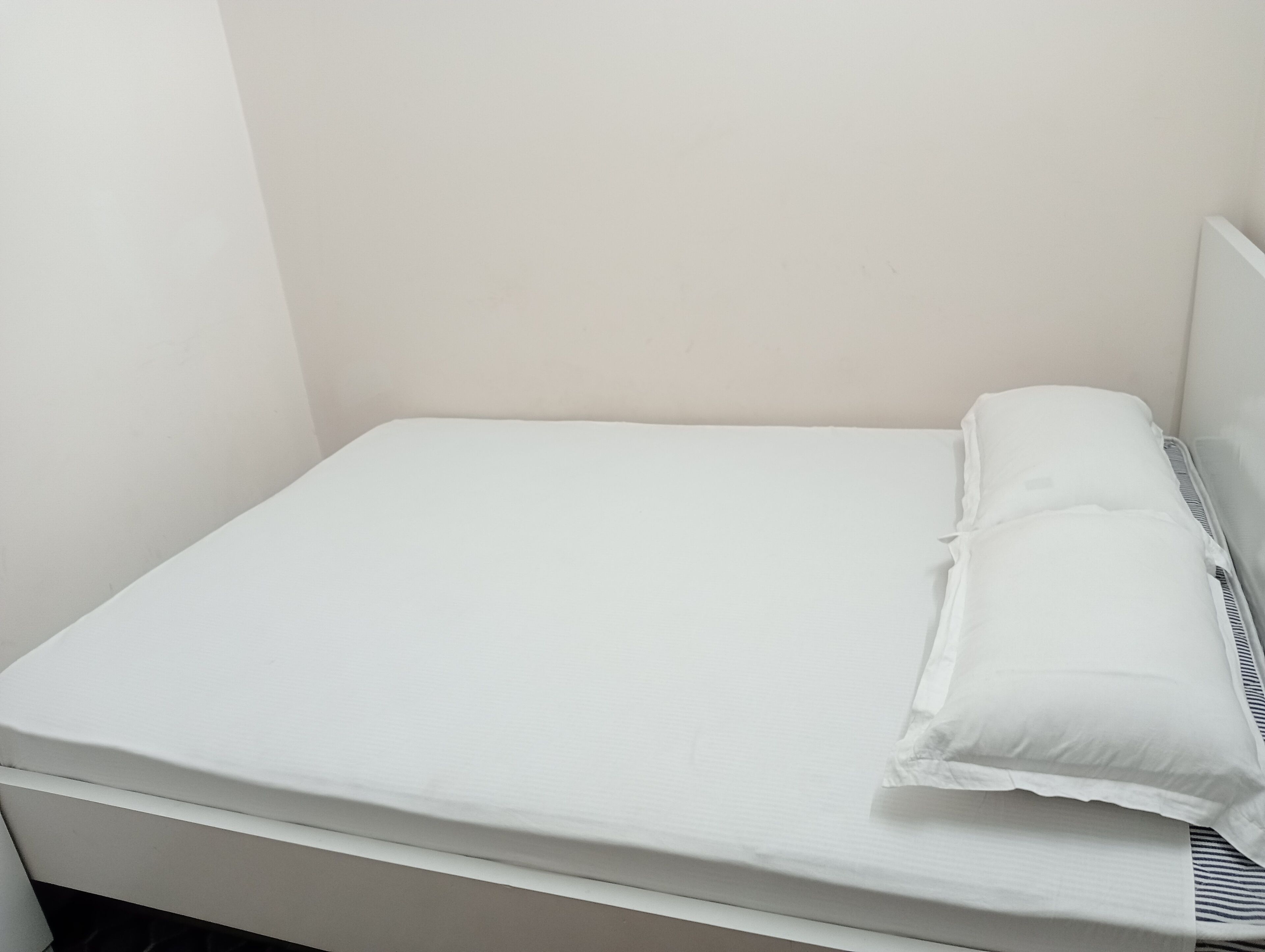 A small, single-bed room with a plain white mattress and pillows, minimal furniture, and a plain wall.