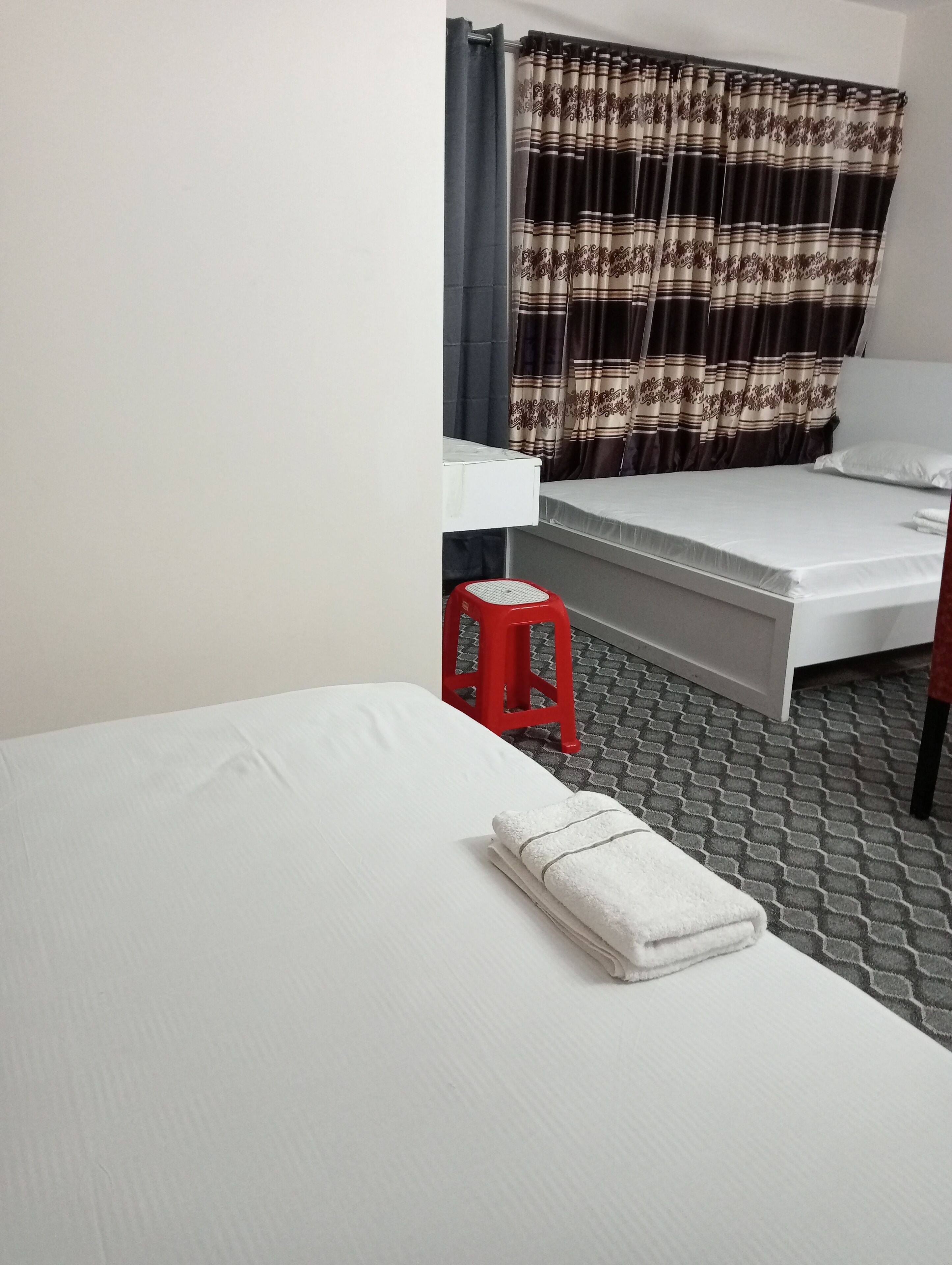 Una cama individual con un colchón blanco y una toalla blanca doblada cuidadosamente. Un taburete rojo está colocado junto a la cama. La habitación tiene una cortina estampada y una pared blanca.