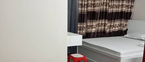 A single bed with a white mattress and a neatly folded white towel. A red stool is placed beside the bed. The room has a patterned curtain and a white wall.