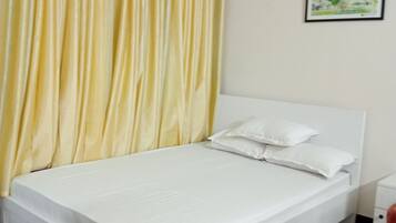 Uma cama de solteiro com roupa de cama branca, uma cortina amarela, um quadro na parede e um tapete estampado no chão.